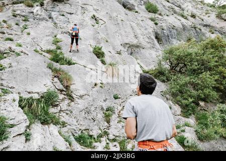 Rear view of male hiker looking at female partner climbing on rocky cliff Stock Photo