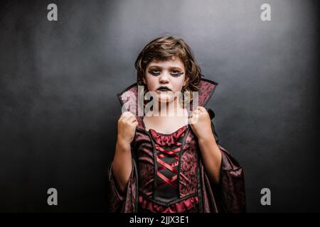 Creepy unemotional little girl with dark makeup in enchantress costume looking at camera against black background during Halloween party Stock Photo