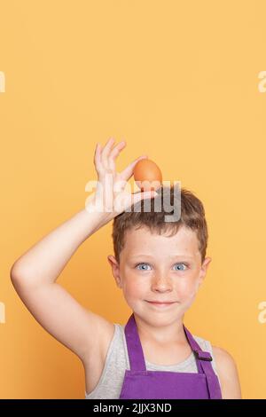 Cute boy in purple chef apron holding egg on head looking at camera while standing against yellow background in light studio Stock Photo