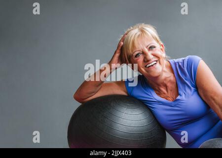 Smiling fit senior Woman leaning on fit ball looking away at gym Stock Photo