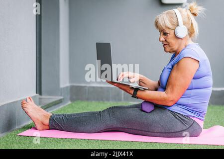 Full body side view of active senior Woman using laptop sitting on yoga mat at gym Stock Photo