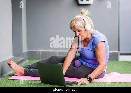 Full body side view of active senior Woman using laptop sitting on yoga mat at gym Stock Photo