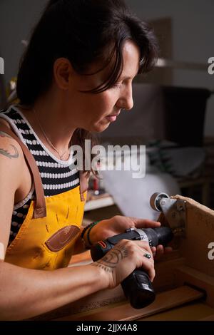 Side view of concentrated adult female carpenter with dark hair and tattoos on arm in casual clothes and apron using drill while screwing metal wheels Stock Photo