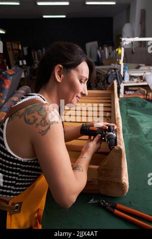 Side view of concentrated adult female carpenter with dark hair and tattoos on arm in casual clothes and apron using drill while screwing metal wheels Stock Photo