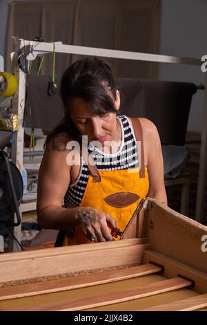 Concentrated adult tattooed female artisan with dark hair in apron using pliers during repairing process at wooden furniture in workshop Stock Photo