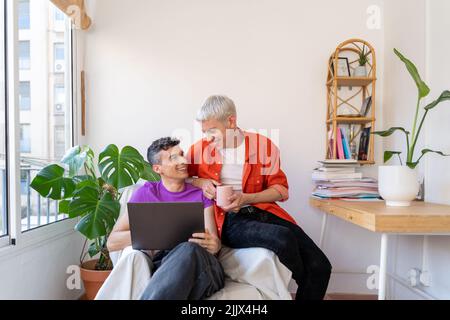 Smiling gay couple using laptop sitting on armchair at home Stock Photo