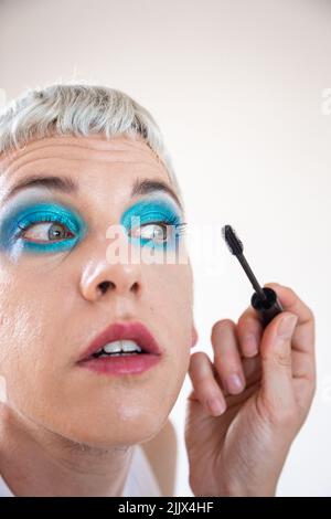 Closeup of transgender man wearing blue eyeshadow applying mascara on white background Stock Photo