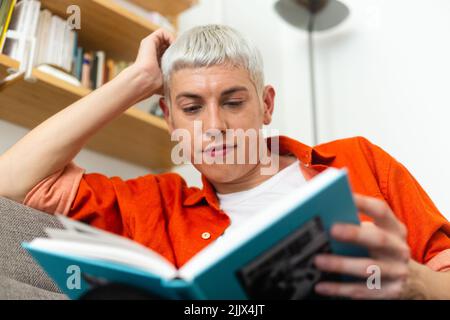 Young androgynous man reading book sitting on sofa at home Stock Photo