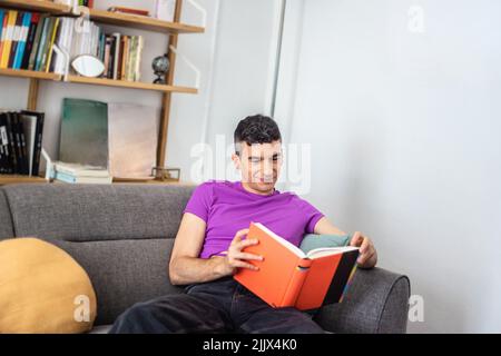 Young androgynous man reading book sitting on armchair at home Stock Photo
