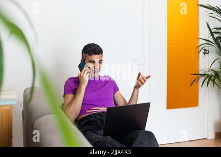 Smiling transgender man using phone and laptop sitting on sofa at home Stock Photo