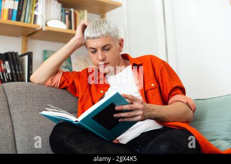 Young androgynous man reading book sitting on sofa at home Stock Photo