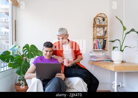Smiling gay couple using laptop sitting on armchair at home Stock Photo