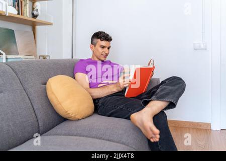 Young androgynous man reading book sitting on armchair at home Stock Photo