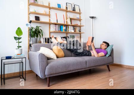 Young androgynous man reading book lying down on armchair at home Stock Photo