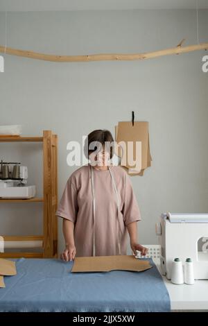 Focused professional dressmaker with measuring tape making patterns on fabric while working at table with special equipment in light atelier Stock Photo