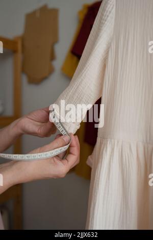 Crop tailor using tape to measure sleeve of handmade white dress while working in studio in daytime Stock Photo