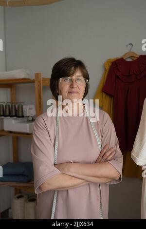 Self assured middle aged Woman with tape measure crossing arms and looking at camera during work in atelier Stock Photo