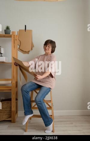 Full body middle aged Woman in casual clothes inspecting paper templates while working in light studio Stock Photo
