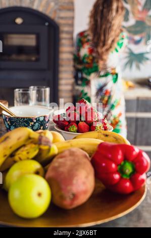 Set of colorful ripe fruits and vegetables placed on table near fruits and dinnerware in light kitchen with faceless Woman Stock Photo