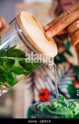 Crop unrecognizable female pouring refreshing water with mint into blender bowl with green kale while preparing smoothie in light kitchen Stock Photo