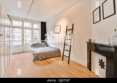 Interior of modern light bedroom furnished with comfortable bed placed on parquet floor near wooden ladder opposite wardrobe near window with curtains Stock Photo