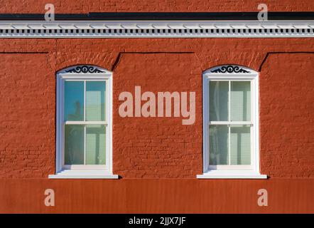 Two white framed windows in a rich colored red brick building with scrollwork and cornice with a brick band at the bottom Stock Photo