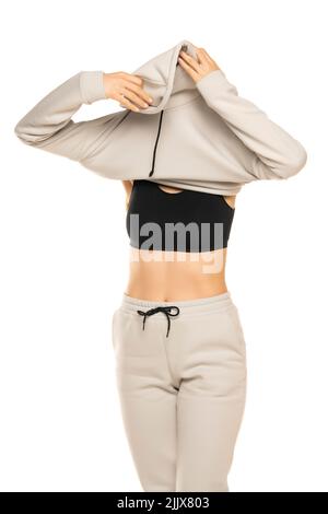 Front view of a young woman takong off her gray tracksuit and hood to a white background in the studio. Stock Photo