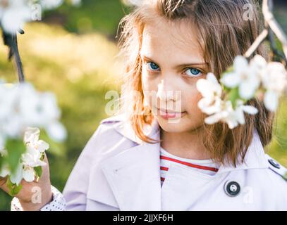 Little girl is walking in spring garder with apple trees and white flowers. Romantic stylish spring kid in coat. Blooming in sunny park. Beautiful nat Stock Photo