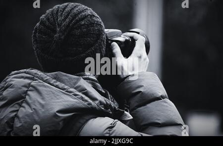 A closeup grayscale back view of a photographer in a hat and winter jacket taking a photo with a camera Stock Photo