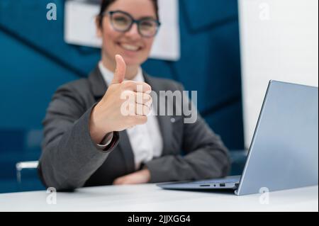 Smiling woman in a business suit shows thumb up while sitting at a desk. Female boss works at a laptop and gestures approval. Stock Photo