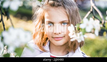 Little girl is walking in spring garder with apple trees and white flowers. Romantic stylish spring kid in coat. Blooming in sunny park. Beautiful nat Stock Photo