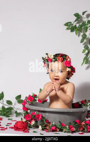 beautiful latina baby girl with brown skin, inside a gray bucket, surrounded by flowers and roses and red, with a white background. little girl suckin Stock Photo