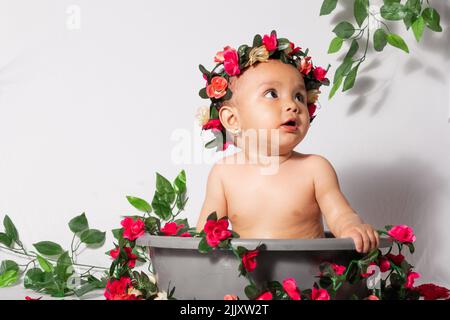 beautiful latina baby girl with brown skin, inside a gray bucket, surrounded by flowers and red roses, with a white background. looking towards the sk Stock Photo