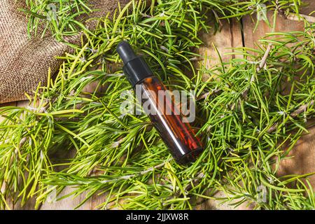 Surrounded by rosemary branches is a brown glass bottle with essential oil capped with a black dropper stopper. Underneath is a sackcloth and wooden p Stock Photo
