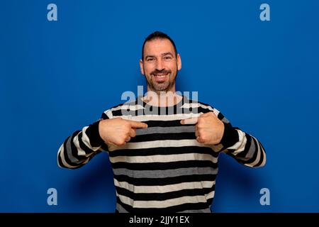 Surprised, amused, smiling, happy caucasian man 40s wear striped pullover looking at camera pointing index fingers on himself isolated on blue Stock Photo