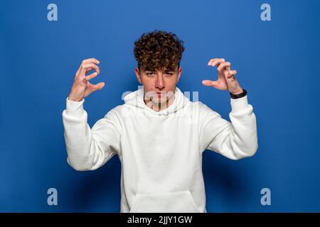 Portrait of a happy young man gesturing with the fingers of his two hands, looking at the camera on a blue background. Stock Photo