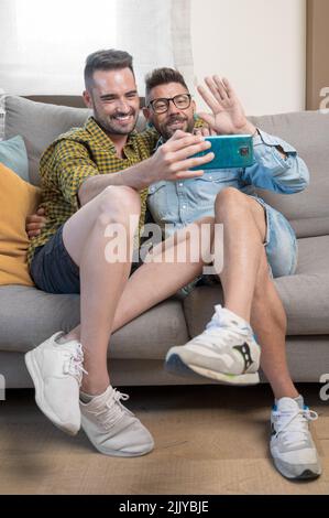 Gay Couple sitting in the sofa At Home Making Video Call On Mobile Phone. High quality photography. Stock Photo