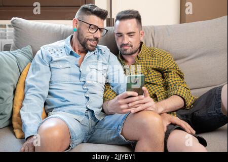Happy young gay couple using mobile phone while sitting on a sofa in the living room. High quality photography. Stock Photo