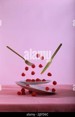 Juicy raspberries on pink plates and berries falling into it , flying knife and fork on pink background with copy space. Stock Photo