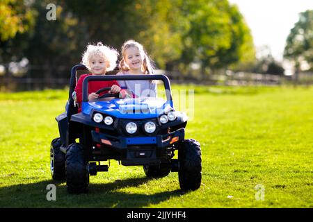 Kids driving electric toy car in summer park. Outdoor toys. Children in battery power vehicle. Little boy and girl riding toy truck in the garden. Stock Photo