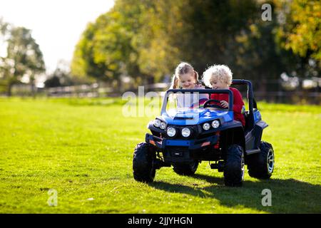 Kids driving electric toy car in summer park. Outdoor toys. Children in battery power vehicle. Little boy and girl riding toy truck in the garden. Stock Photo