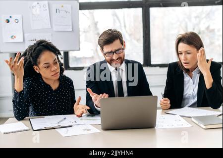 Upset business employees of a different nationalities are sitting at the workplace in a modern office, experience stress of work deadline, disappointed with the outcome of team work, confused Stock Photo