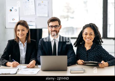 Creative business team in the office. Positive successful smart colleagues, dressed in formal wear, sitting at a desk in their modern office, looking at the camera, smiling friendly Stock Photo