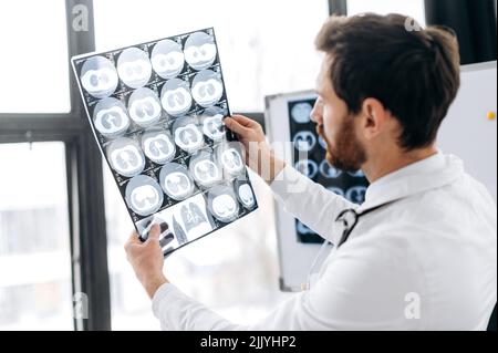 Professional doctor looking at x-ray at his cabinet, caucasian male therapist in white medical uniform and glasses concentrated learning patient's x-ray, standing in his medical office Stock Photo