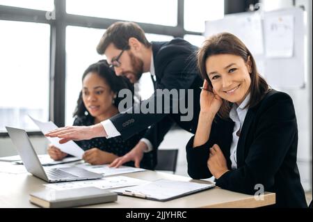 Beautiful confident caucasian woman in a formal wear, ceo company, a financial manager, sitting at a desk with her multiracial colleagues in the office, looking at the camera, smiling friendly Stock Photo