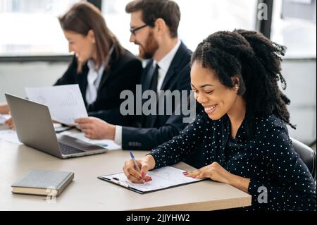 Professional african american young woman in a shirt, a financial manager or ceo, sitting at a desk with her colleagues in the office, concentrated examines the documents, financial report, smiling Stock Photo