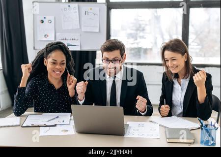 Cheerful creative business partners working together in the office. Multiracial colleagues sitting in modern office, using laptop, they are happy get good deal, big profit. The concept of win, triumph Stock Photo