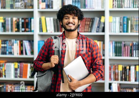 Happy confident indian or arabian male student of university, in stylish casual wear, with backpack and laptop, stands in a library against the background of bookshelves, looks to the camera, smiles Stock Photo