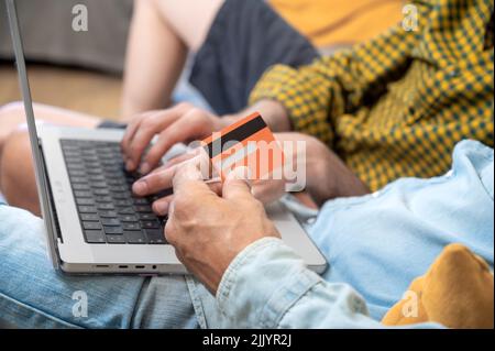 Close up of an unrecognizable gay couple using laptop computer while sitting on a couch at home, shopping online with a credit card. High quality photography. Stock Photo