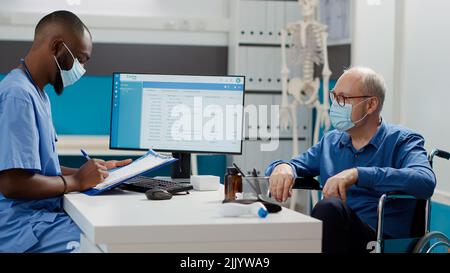 Medical worker taking notes at consultation with senior man suffering from physical disability. Consulting male wheelchair user with impairment during coronavirus pandemic, checkup visit. Stock Photo